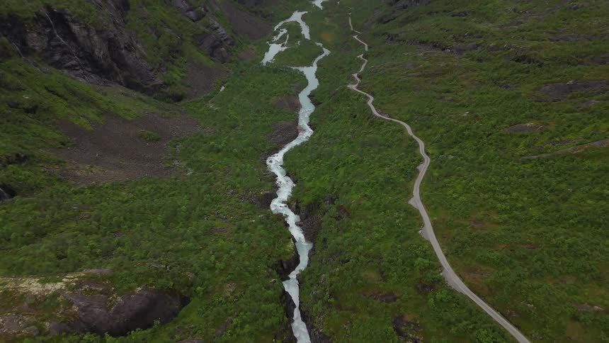 An epic aerial reveal shot flies backward over a turquoise river, unveiling a stunning green mountain valley with a winding road