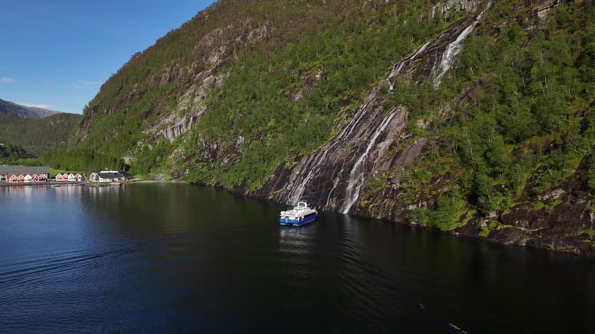 A majestic aerial view of a tourist ferry sailing past a beautiful waterfall in a Norwegian fjord