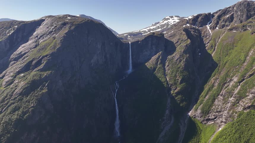 A stunning aerial view pulls back to reveal a majestic waterfall cascading down a dramatic, green mountain valley under a clear blue sky