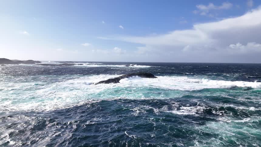 A dramatic slow-motion shot shows turbulent ocean waves crashing over jagged rocks on a windy day