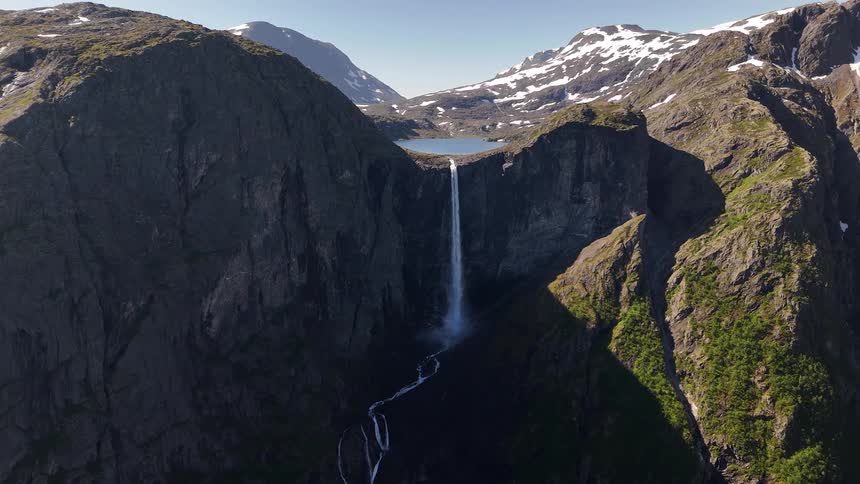 A stunning static aerial shot captures a tall waterfall originating from a high-altitude lake and cascading down a sheer cliff into a rugged mountain valley