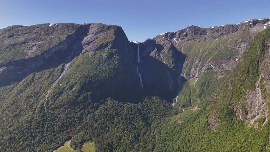 A static wide aerial shot shows the majestic Mardalsfossen waterfall flowing down a vast mountain range covered in green forests under a clear summer sky in Norway