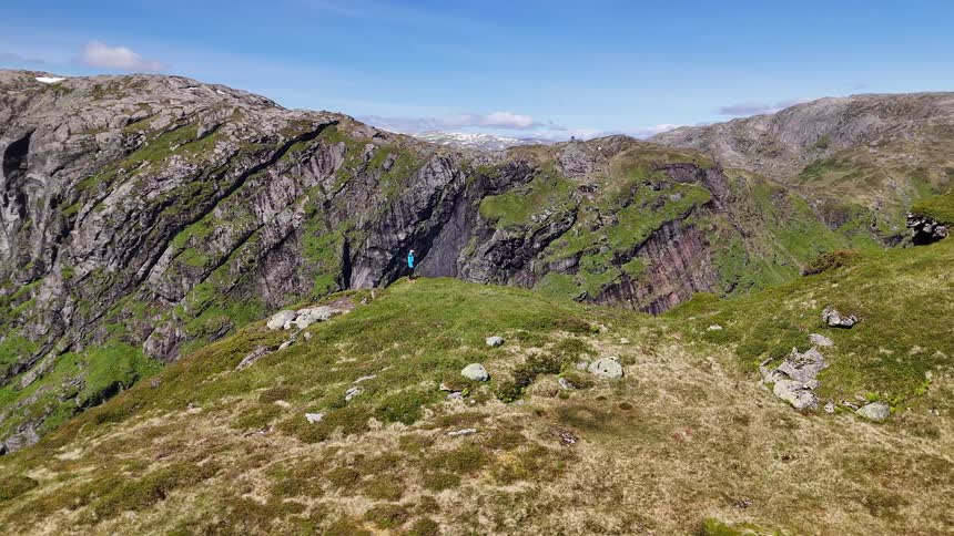 A slow aerial pull-back reveals a lone hiker standing on a dramatic, green mountain ridge, taking in the immense, rugged landscape on a clear day