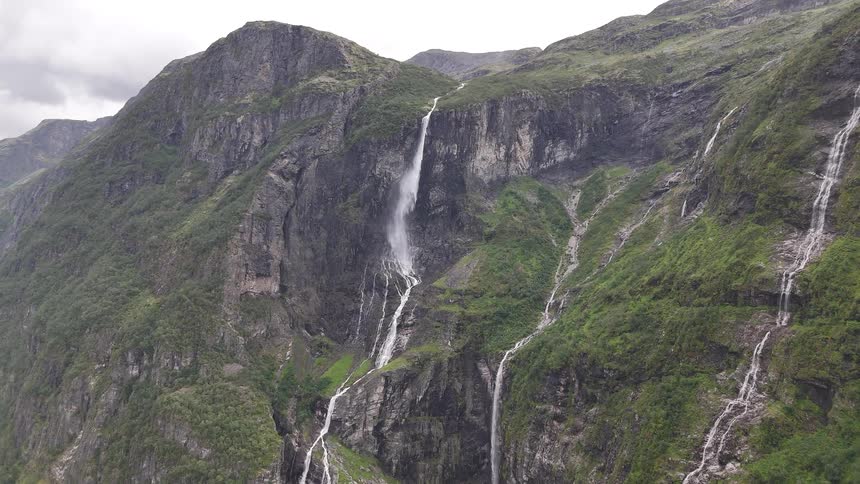 Multiple powerful waterfalls cascade down the steep, rocky cliffs of a lush green mountain on an overcast day