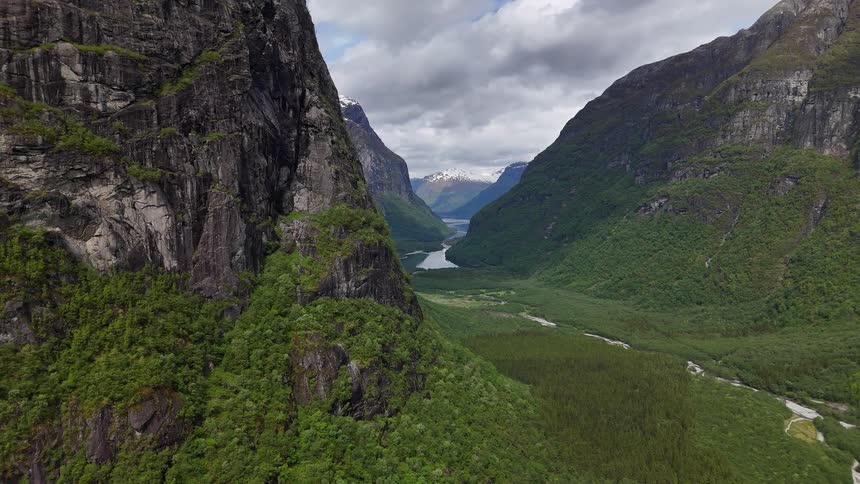 A stunning aerial view slowly reveals a majestic green valley between towering rocky mountains, with a river leading towards snow-capped peaks in the distance