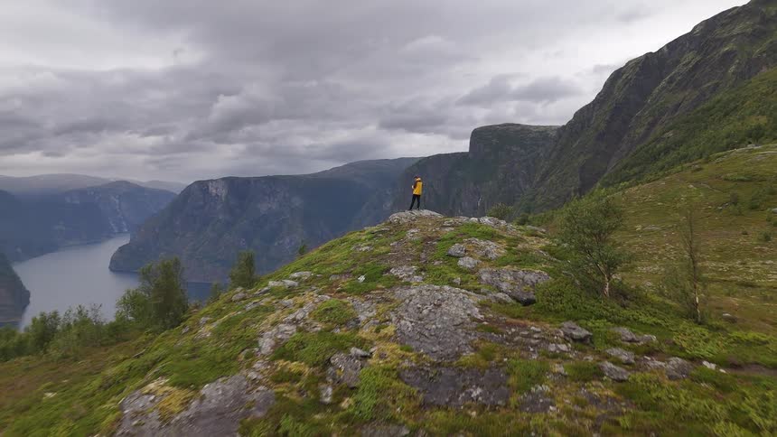 An aerial shot flies towards a hiker in a yellow jacket, revealing a stunning view of a Norwegian fjord from a mountain overlook on a cloudy day