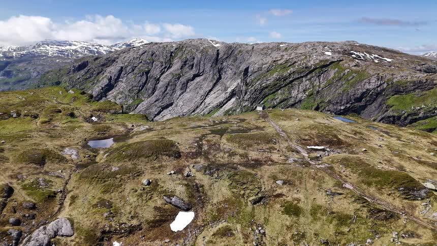 An epic aerial pullback shot over a rugged mountain plateau reveals a stunning fjord and snow-capped peaks in the distance