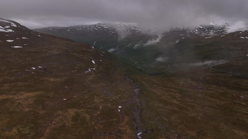 A vast aerial view shows a moody mountain valley with a river running through it, as heavy clouds and mist hang over the snow-capped peaks