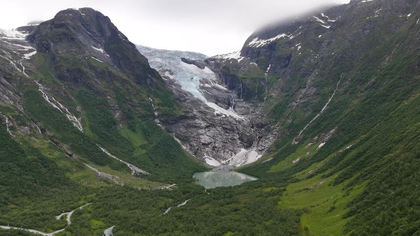 A stunning aerial panorama reveals a majestic glacier and its meltwater lake, surrounded by towering green mountains and cascading waterfalls in Norway