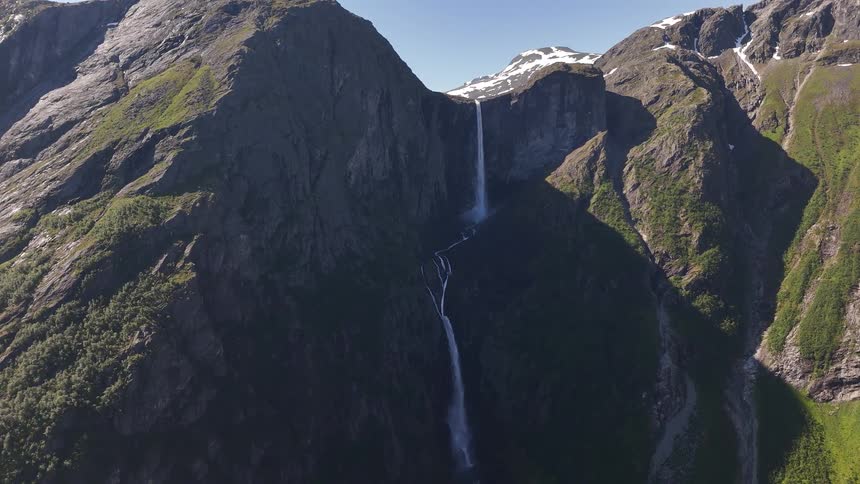 A breathtaking static aerial shot captures a tall waterfall flowing down a majestic, green mountain under a clear blue sky