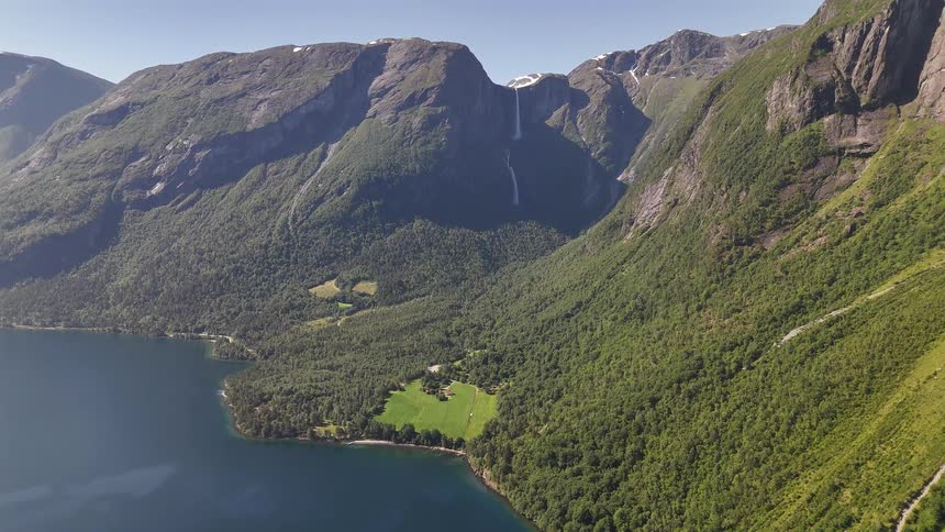 Epic aerial pullback shot reveals a majestic waterfall cascading into a serene fjord surrounded by lush green mountains in Norway