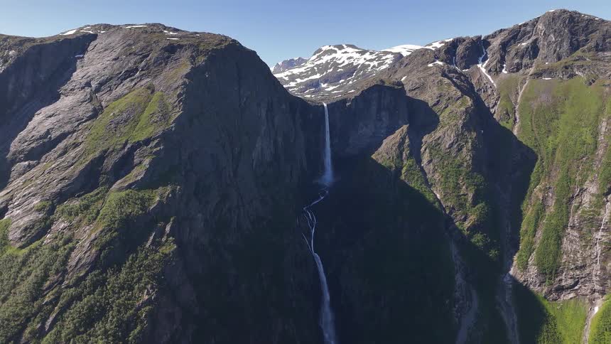 A slow aerial pullback reveals a majestic waterfall flowing from a snow-capped mountain range down a rugged cliffside