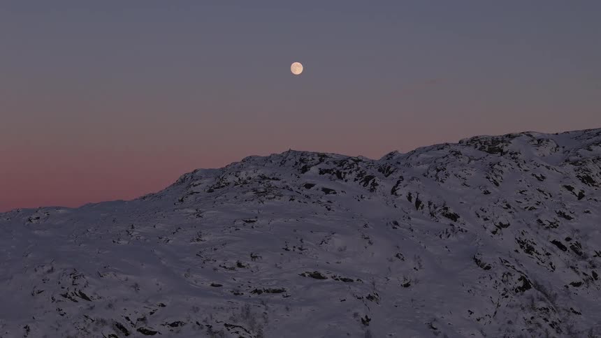 A serene time-lapse captures the full moon rising over a vast, snowy mountain landscape against a beautiful twilight sky