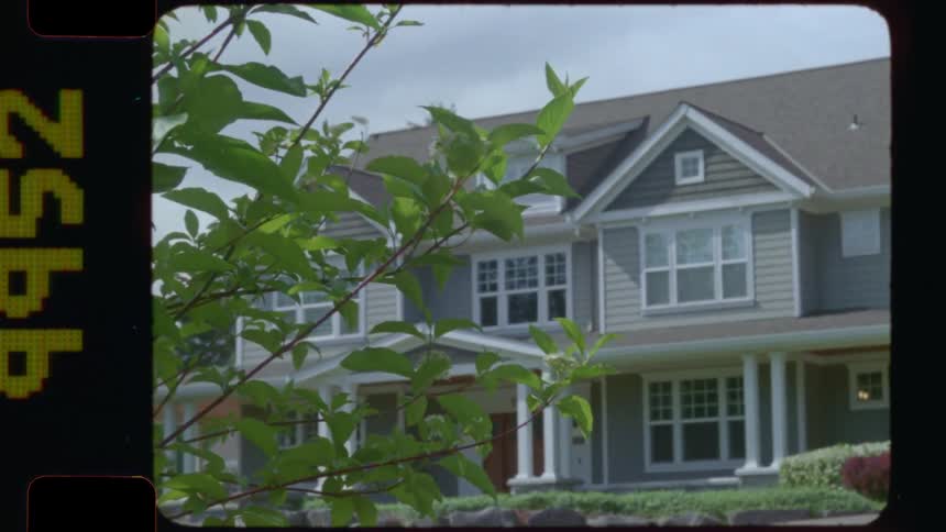 A quiet suburban house is viewed from behind green tree leaves in a vintage home movie style 16mm film shot