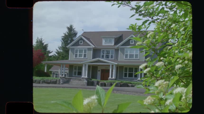 A vintage 16mm film shot of a classic suburban two-story house with a green lawn, framed by blooming flowers