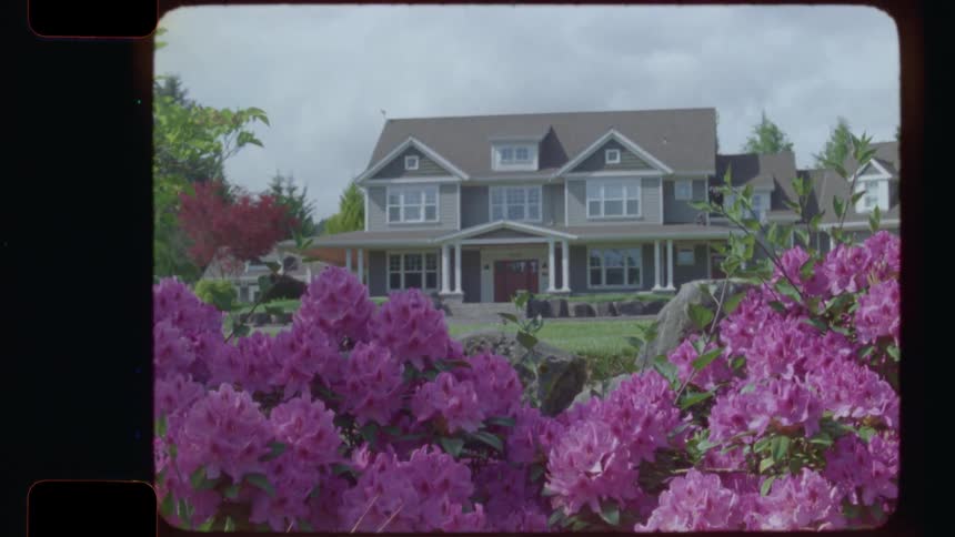 A slow reveal from behind vibrant pink rhododendron flowers shows a large, idyllic suburban house on a sunny day, captured on vintage 16mm film