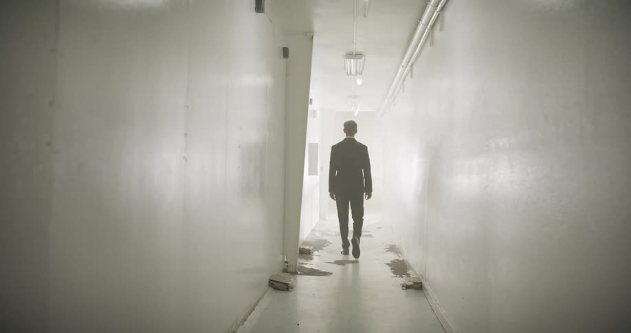 A mysterious businessman in a dark suit walks slowly down a long, derelict white hallway and disappears into a thick white fog
