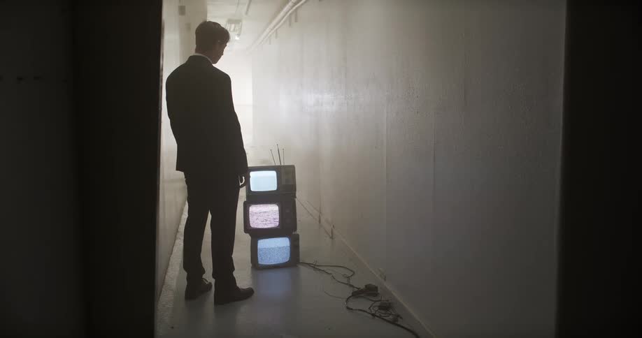 A mysterious man in a suit slowly walks down a long, foggy hallway towards a stack of old televisions displaying static