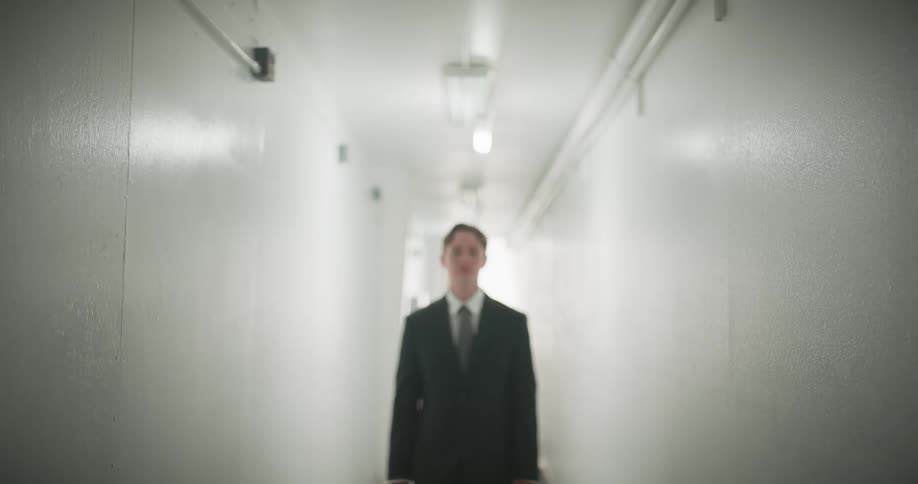 A young man in a black suit walks down a long, white hallway, coming into focus as he approaches the camera