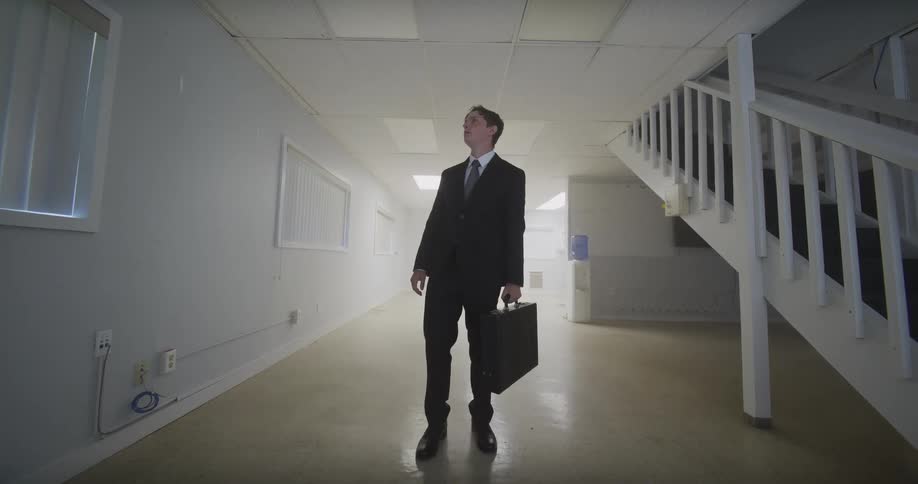 A businessman in a suit walks through a sterile, empty basement office space, looking around at the vacant room