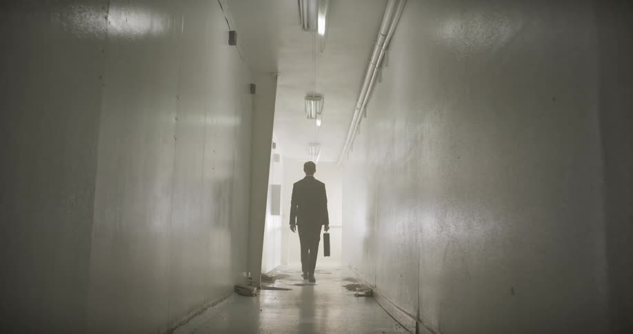 A mysterious businessman walks away down a long, dark, and decrepit hallway into a bright, foggy light