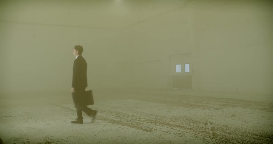 Businessman with a briefcase walks in slow motion through a large, desolate, and dusty warehouse