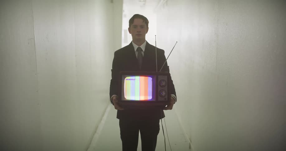 A slow tilt-up reveals a young man in a formal suit holding a vintage television displaying a color bar test pattern in a long, hazy hallway