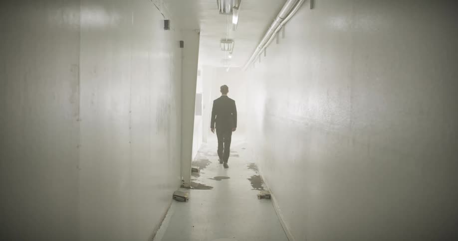 A mysterious man in a suit walks away down a stark white corridor and vanishes into a cloud of smoke