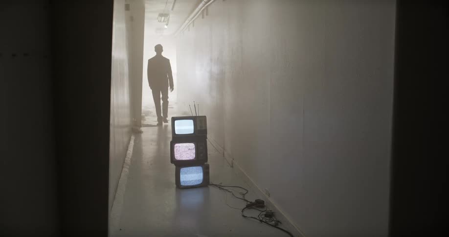 A man in a suit walks away from a stack of vintage televisions displaying static in a long, foggy hallway