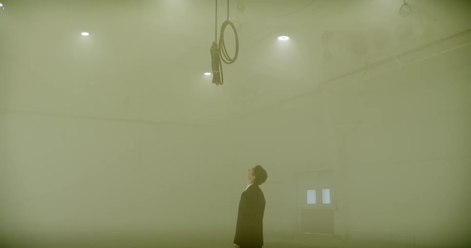 Camera tilts down in a foggy, empty warehouse to reveal a businessman with a briefcase looking up at a hanging noose