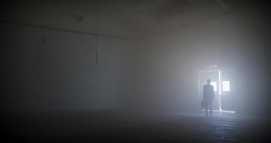 A businessman carrying a briefcase walks through a dark, foggy warehouse and exits through a brightly lit doorway