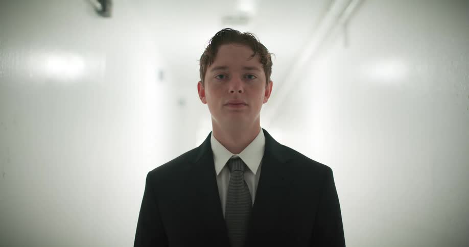 A serious young man in a business suit stands still in a stark white hallway as the lights flicker dramatically
