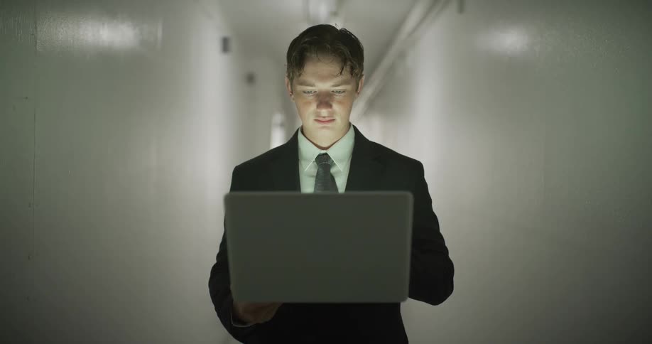 A focused young businessman in a suit walks through a sterile hallway, his face illuminated by his glowing laptop screen