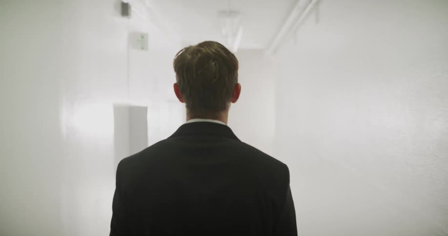 A man in a suit walks down a long, stark hallway, journeying from darkness into a bright, hazy light