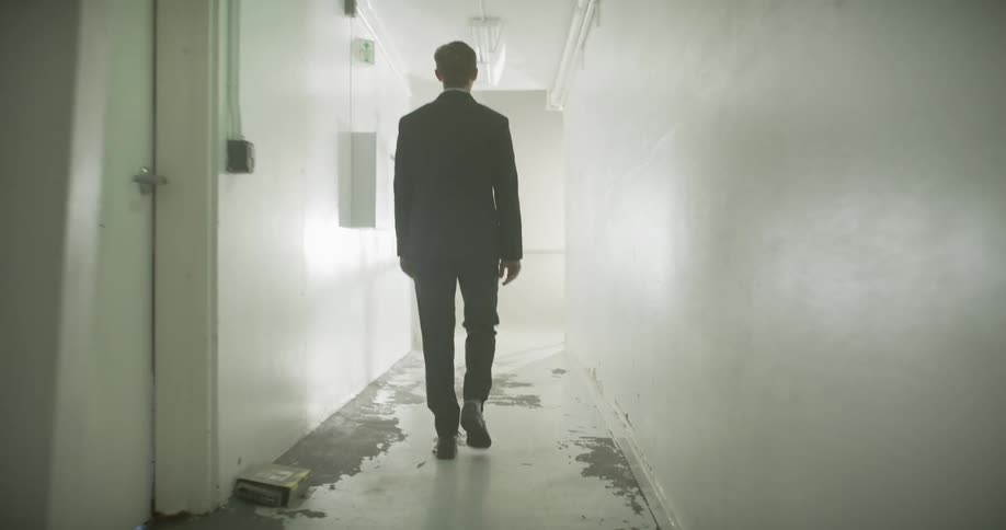 A mysterious man in a black suit walks in slow motion down a long, gritty hallway towards a bright, smoky light