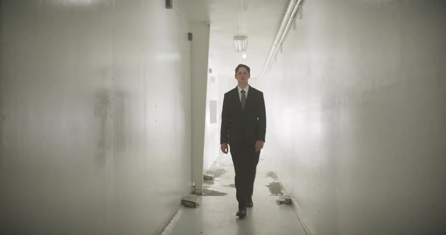 A mysterious man in a dark suit confidently walks down a long, foggy, and dilapidated white hallway towards the camera