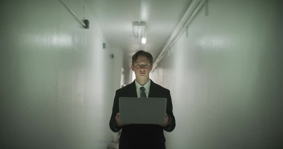 An intense young man in a suit stares directly into the camera while holding an open laptop in a stark hallway with flickering lights