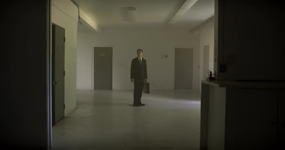 A mysterious businessman in a suit carries a briefcase through an empty, stark white institutional room