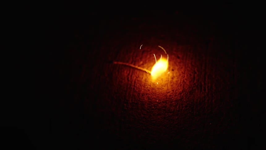 A close-up shot captures a matchstick igniting and burning in slow motion on a dark, textured surface, emitting a bright flame and sparks