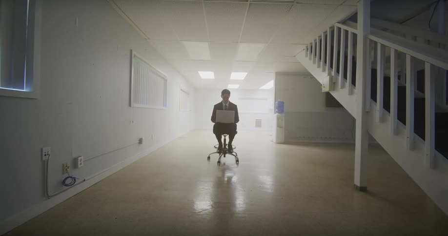 A lone businessman in a suit works on his laptop while sitting in a chair in the middle of a vast, empty, white office space