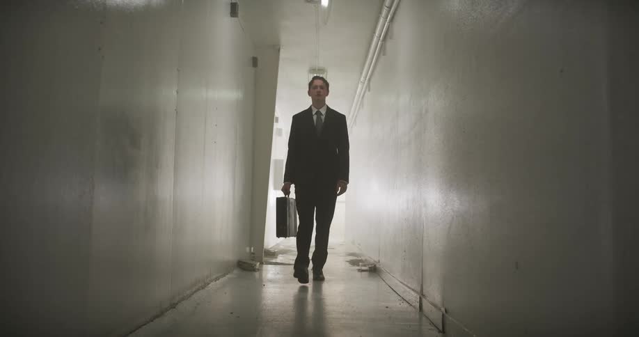 A businessman carrying a briefcase walks with purpose down a long, dark, and decrepit hallway towards the camera in slow motion