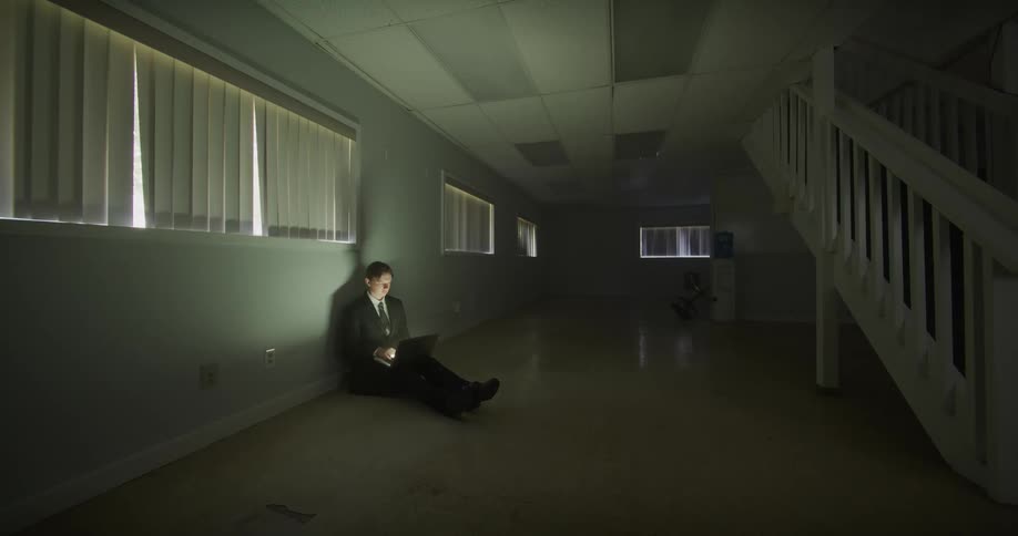 Illuminated by his laptop screen, a businessman sits alone on the floor of a dark, empty office