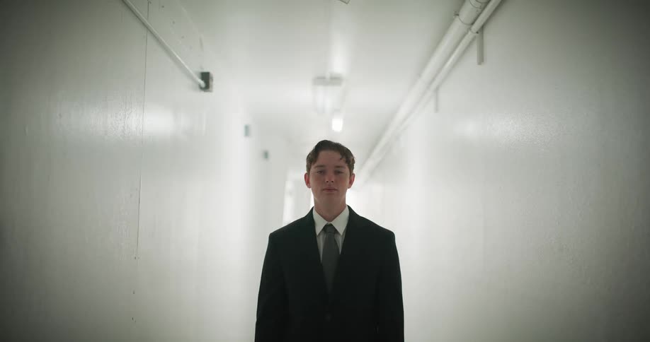 A young man in a suit stands still in a stark white hallway as the overhead lights flicker on and off