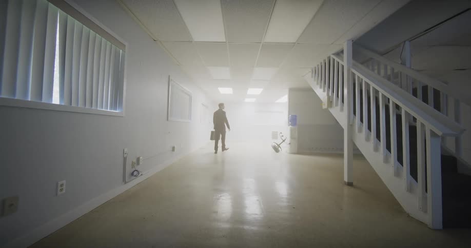 A man in a business suit carrying a briefcase walks through an empty, sterile basement office and disappears into a bright, ethereal light