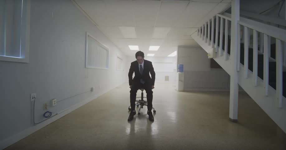 A man in a business suit walks into a stark white empty room and sits on a single rolling stool in the center
