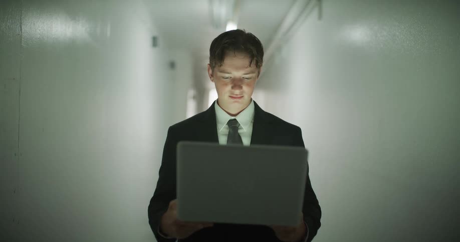 A focused young businessman in a suit walks through a stark, flickering hallway, his face lit up by the glow of his laptop