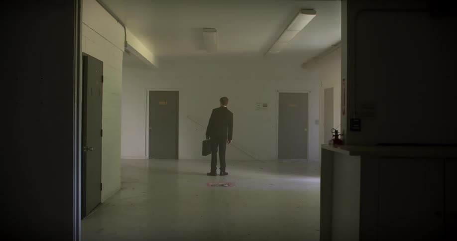 A mysterious businessman in a suit carries a briefcase through a hazy, abandoned building before choosing a door