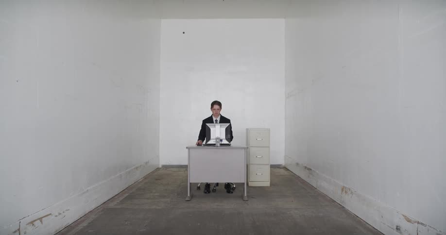 A businessman in a suit enters a stark, empty white room and sits down at his minimalist desk to begin working