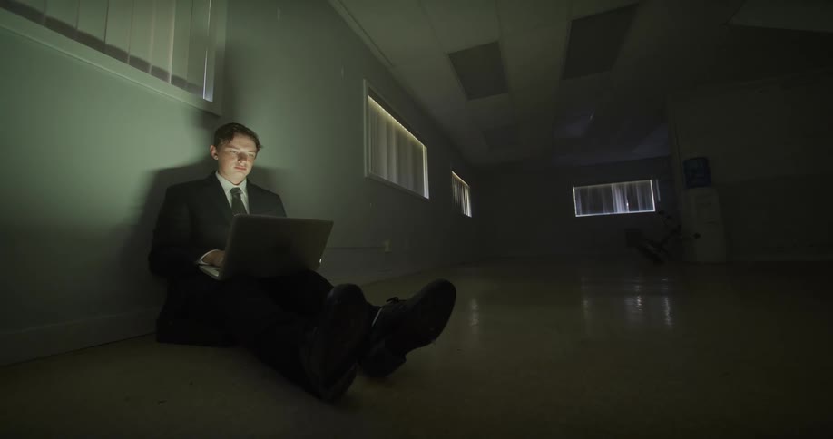 A businessman in a suit sits alone on the floor of a dark, empty office, intensely focused on his work on a laptop