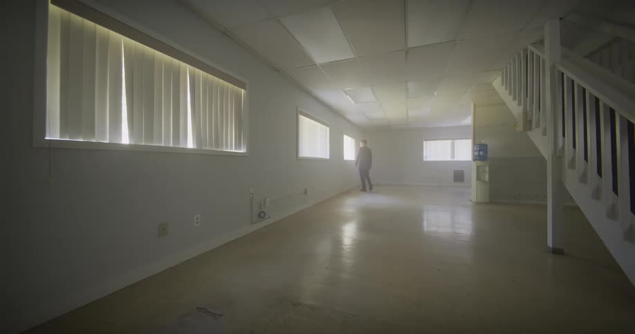 A man in a black suit walks slowly through a long, empty, and hazy room, creating a sense of mystery and solitude